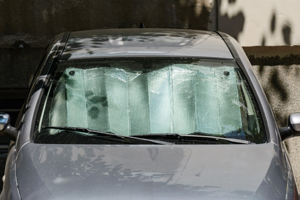 Foil sun shades under the windshield of a gray car parked outdoors on a sunny summer day. Reflective sun shield made of metallic silver foil protects vehicles from direct sunlight. Front view.